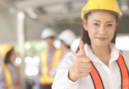 Close up image of confident Asian Female architect or engineer  holding blue print paper, standing in front of her team smiling , looking at camera and showing her thumb up.Selective focus on woman's thumb.の写真素材