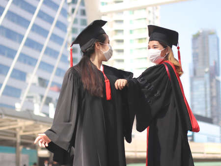 Two graduated women in graduation gowns and medical face masks,  elbows greeting to avoid the spread of virus.. Graduation Class of 2020. Covid-19 prevention and new normal concept.の写真素材