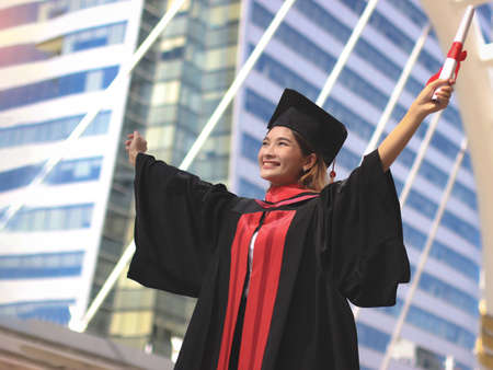 Happy Asian  graduated women in graduation gowns , holding diploma, smiling and open arms.. City building background. Education, successful concept.の写真素材