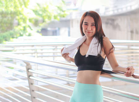 Happy attractive Asian sportswoman in sportswear standing against bridge fence, smiling and looking at camera.の写真素材