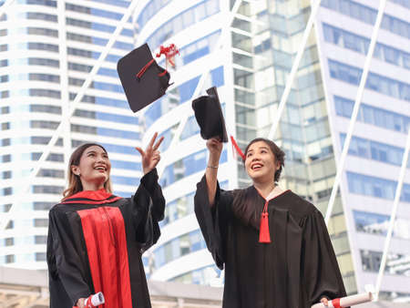 Two happy graduated women in graduation gowns, throwing  hats up and smiling happily. City building background. Education, Friendship concept.の写真素材