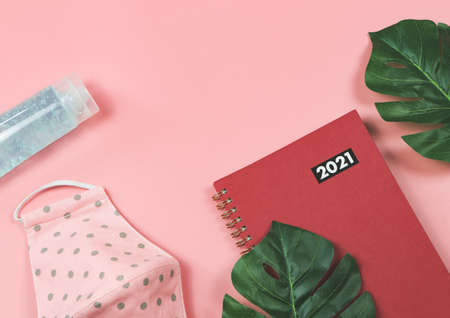 Top view or flat lay of red diary or planner 2021 with monstera leaves, fabric  face mask, alcohol sanitizer gel on pink background. New year 2021 planning, Covid-19 prevention, new normal concept.の写真素材