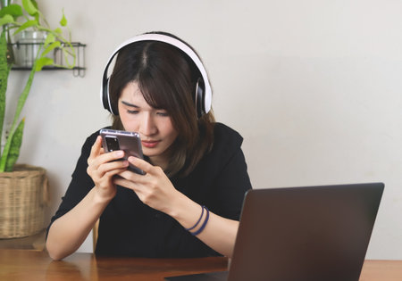 Asian woman in black shirt wearing headphones, sitting at wooden table with computer laptop, using mobile phone. white wall with house plant background.の写真素材