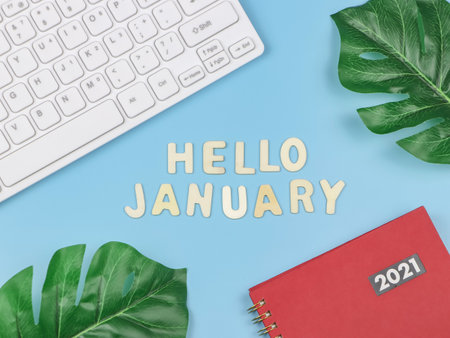 Wooden letters HELLO JANUARY  with red 2021 diary or planner, computer keyboard and monstera leaves...
