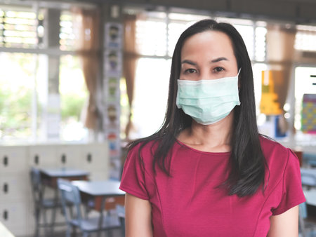 Portrait of Asian female teacher wearing protective face mask, standing in empty classroom,  looking at camera.の写真素材