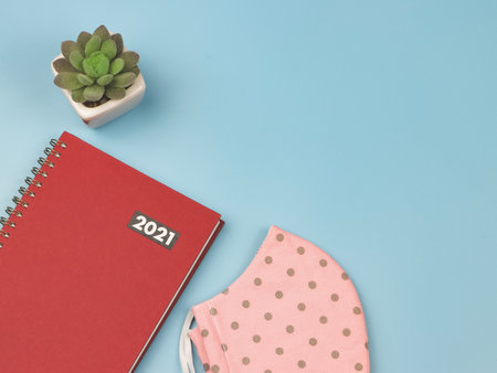 Top view or flat lay  of red diary or planner 2021 with pink polka dot fabric mask plant pot on blue background. New year 2021 planning, COVID19 prevention and new normal concept.の写真素材