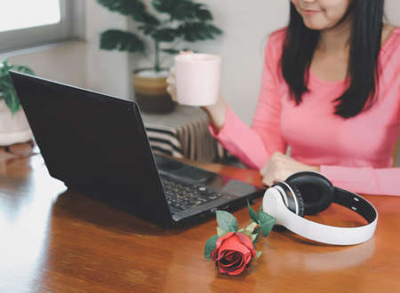 Cropped photo of woman holding cup of coffee, sitting at table with computer laptop, headphones and...
