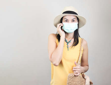 Portrait of asian female tourist wearing yellow sleeveless shirt, hat and medical protective mask, holding woven bag, smiling and looking at camera. Covid19 prevention while traveling.の写真素材