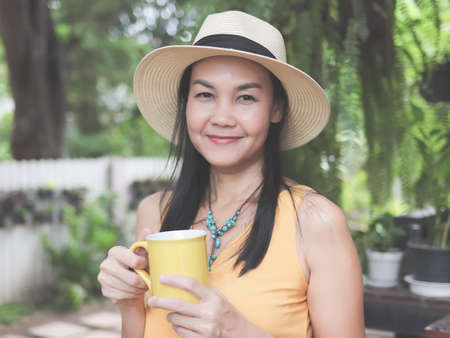 Portrait of Asian woman  wearing yellow sleeveless shirt, hat holding yellow coffee cup, standing in...