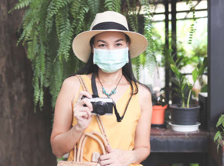 Portrait of Asian female tourist wearing yellow sleeveless shirt, hat and fabric  protective mask, holding camera, standing in the garden. prevention while traveling.の写真素材
