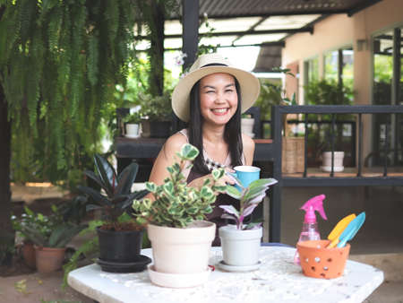 Portrait of happy Asian woman wearing hat and sleeveless shirt sitting in the garden holding blue cup of coffee,  smiling happily and looking at camera.の写真素材