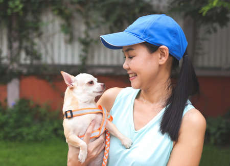 Portrait of Asian woman wearing blue sportswear, standing in the garden, holding brown Chihuahua dog with leash, smiling and looking each other.の写真素材