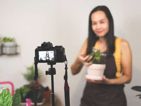 Asian female gardener in  sleeveless shirt and apron, using camera to record video about houseplant  taking care. Asian woman selling houseplant online. Selective focus.の写真素材