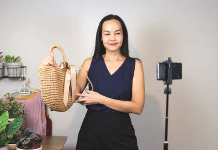 Asian woman  working at home,  using mobile phone to live or record video selling her woven bag. Indoor,  selective focus. Business and online selling concept.の写真素材