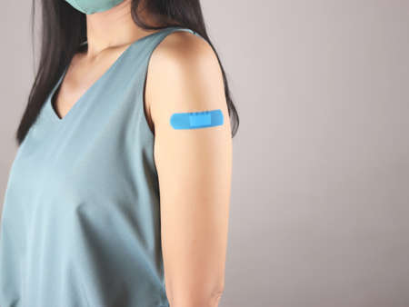 Cropped image of vaccinated Asian woman in green sleeveless t-shirt and cloth face mask  with Plaster Bandage On her Arm After Coronavirus vaccine injection. Isolated on gray background.の写真素材
