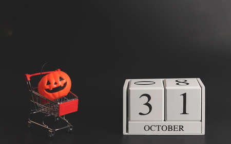 Front view of wooden calendar 31 October with plastic Halloween pumpkin on shopping cart or trolley on black background.の写真素材