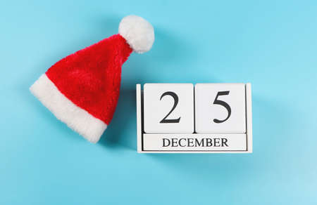 Top view or flat lay of wooden calendar December 25 with bright red Santa Claus hat on blue...
