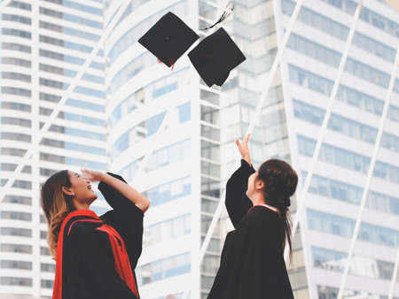 Portrait of Two happy graduated women in graduation gowns, throwing  hats up and smiling happily. City building background. Education, Friendship concept.の写真素材