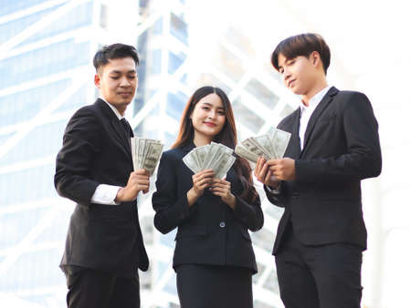 Portrait of successful Asian business woman and two businessmen in suit standing  and holding fan of money in dollar banknotes, smiling and looking at camera, city building background.の写真素材
