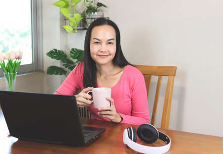 Portrait of Asian woman sitting with computer notebook, holding cup of coffee, smiling and looking...