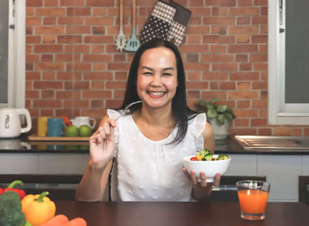 Portrait of Asian woman eating salad in the kitchen, smiling and looking at camera. Healthy lifestyle concept.の写真素材