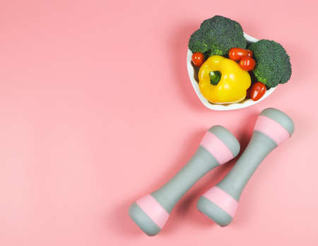 Top view of vegetables  capsicum, broccoli and tomato in heart shape plate and pink dumbbells on...