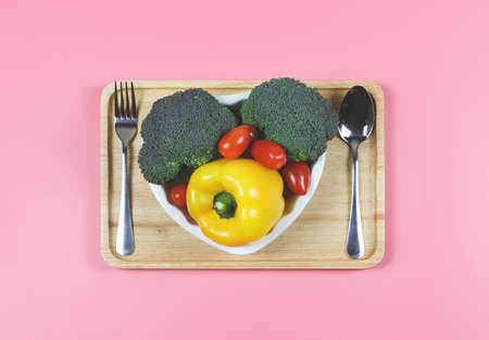 Top view or flat lay of vegetables  capsicum, broccoli and tomato in heart shape plate with fork and spoon in wooden tray  on pink background with copy space, healthy lifestyle and weight loss concept.の写真素材