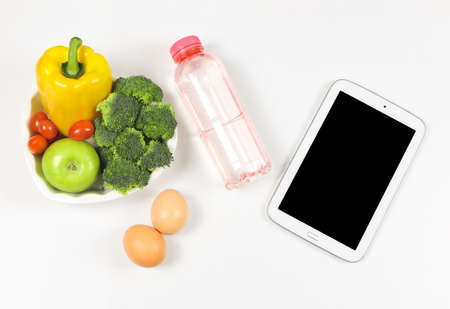 Top view or flat lay of black blank screen computer tablet , vegetables and fruit in heart shape plate, bottle of water and two eggs on white background. Healthy food.の写真素材
