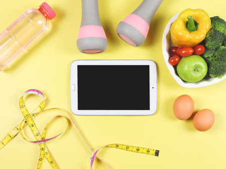 Top view or black blank screen computer tablet , vegetables and fruit in heart shape plate, bottle of water, measuring tape,  pink dumbbells and eggs on yellow background.の写真素材