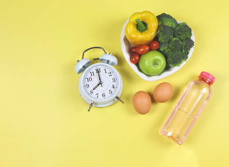 Top view of vegetables  and fruit  in heart shape plate  with white vintage alarm clock, eggs  and pink bottle of water on yellow background. healthy lifestyle  and intermitten fasting concept.の写真素材