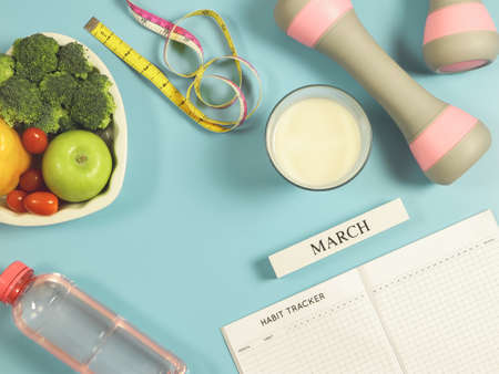 Top view of habit tracker book, wooden calendar March, gray pink dumbbells, measuring tape, water, milk  and vegetables in heart shape plate  on blue background with copy space. Healthy food, lifestyle concept.の写真素材