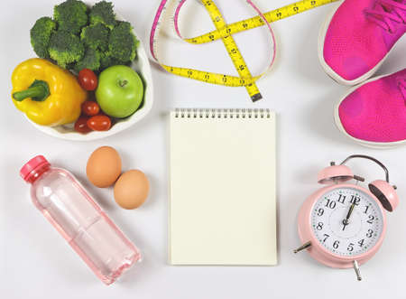 Top view of open notebook, vegetables and fruit in heart shape plate, bottle of water, measuring tape,  pink alarm clock, eggs and pink sneakers on white background.の写真素材