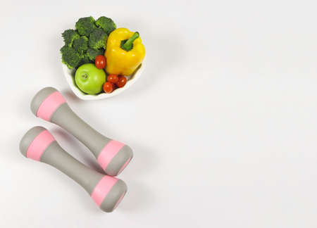 Top view of vegetables  capsicum, broccoli, tomatoes and green apple in heart shape plate and pink dumbbells on white background with copy space. Healthy lifestyle concept.の写真素材