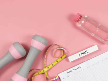 Top view or flat lay of habit tracker book, wooden calendar April, gray pink dumbbells, bottle of water  and measuring tape on pink background with copy space. Self development and active lifestyle concept.の写真素材