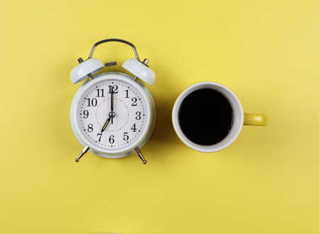 Top view or flat lay of cup  of black coffee and white vintage alarm clock on yellow background.