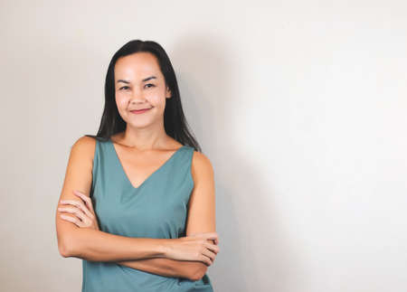 portrait of Asian woman with long back hair wearing green sleeveless blouse, standing with arms crossed, smiiling and looking at camera on white background with copy space.の写真素材