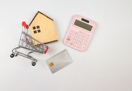 Top view or flat layout of wooden house model in shopping trolley, pink calculator and credit card on white background, home purchase calculation concept.の写真素材