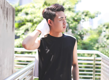 Portrait of Asian young man wearing sportswear, standing and wiping sweat with towel outdoor on...