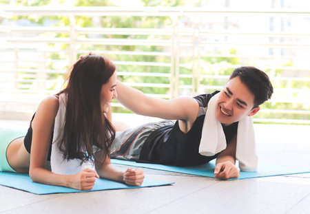Portrait of Asian happy woman and man in yoga sport outfit, practicing yoga, lying down on yoga mat, smiling and a man touch woman's head. Terrace background.の写真素材