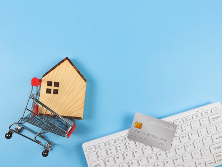 Top view or flat layout of wooden house model in shopping trolley, computer keyboard and credit card on blue background with copy space, home purchase concept.の写真素材
