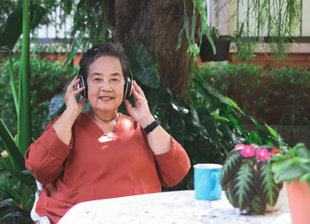 Portrait of happy and healthy  Asian elderly  woman sitting with coffee cup and  flower pots in outdoor  garden, listening to favorite music from headphones, smiling and looking at camera.の写真素材