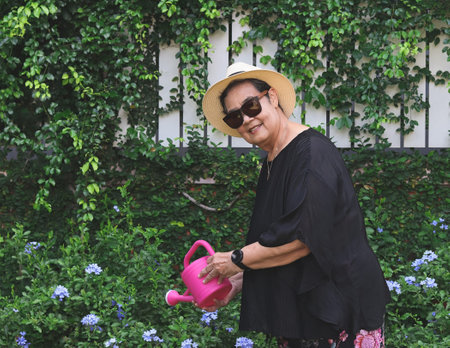 Portrait of happy and healthy senior asian woman wearing black blouse, sunglasses and hat, holding pink watering can watering plants  in the garden, smiling and looking at camera.の写真素材
