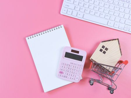 Top view or flat layout of wooden house model in shopping trolley, pink calculator, blank page notebook  and computer keyboard  on pink  background with copy space, home purchase concept.の写真素材