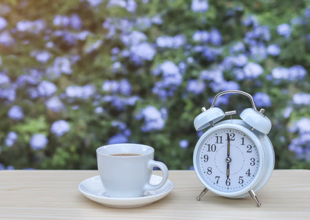 Front view of white vintage alarm clock and cup of coffee on the table in the garden with purple...