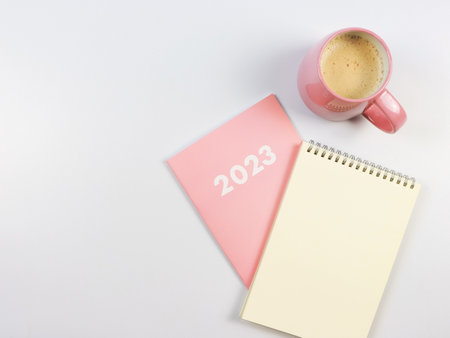 Top view or flat lay of blank paper note book on pink diary 2023 and pink cup of coffee on white...