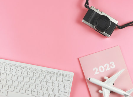 Top view or flat lay of white computer keyboard, pink diary 2023, airplane model  and camera on pink...