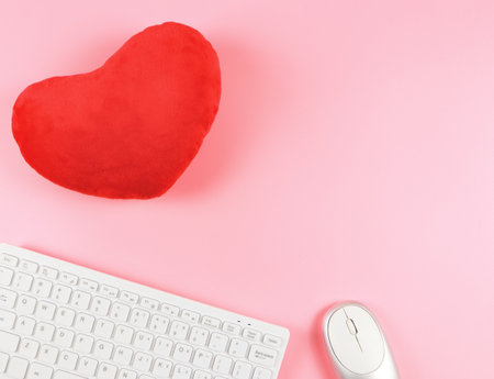 Top view or flat lay of computer keyboard and mouse with red heart shape pillow on pink background.の写真素材