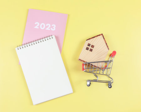 Top view or flat layout of wooden house model in shopping trolley or shopping cart  with   blank page notebook and pink diary or  2023   on yellow  background with copy space, home purchase plan  concept.の写真素材