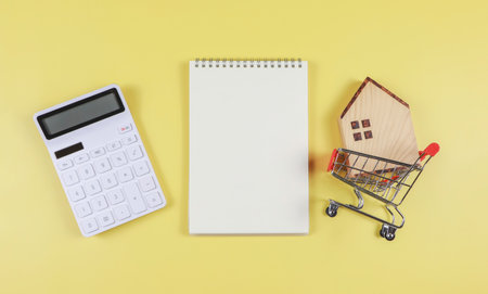 Top view or flat layout of wooden house model in shopping trolley, white calculator, blank page opened  notebook  on yellow  background with copy space, home purchase concept.の写真素材
