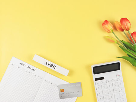 Top view or flat layout of Habit tracker book,  white calculator, credit card, wooden calendar April and tulips on yellow background.の写真素材